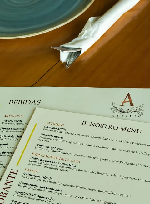 La carta impresa de Attilio, con la sección Il nostro menu y la de bebidas, sobre una mesa de madera junto a un plato turquesa