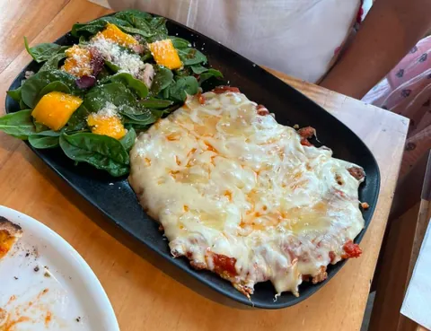 Milanesa napolitana gratinada con tres quesos sobre salsa de tomate, junto a una ensalada de espinaca con mango, en un plato negro